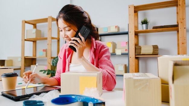 Młoda bizneswoman Azji przy użyciu telefonu komórkowego, odbieranie zamówienia i sprawdzanie towaru na stanie, praca w domowym biurze. Właściciel małej firmy, dostawa na rynek online, koncepcja niezależnego stylu życia.
