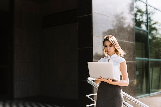 Bezpłatne zdjęcie młoda bizneswoman pozycja na zewnątrz biurowego używa laptopu