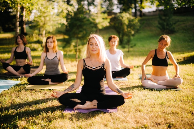 Młoda blond kobieta prowadzi zajęcia jogi w parku przyrody w słoneczny letni poranek. trenerka fitness jogi prowadzi swoją grupę do relaksującej medytacji ze światłem słonecznym za sobą