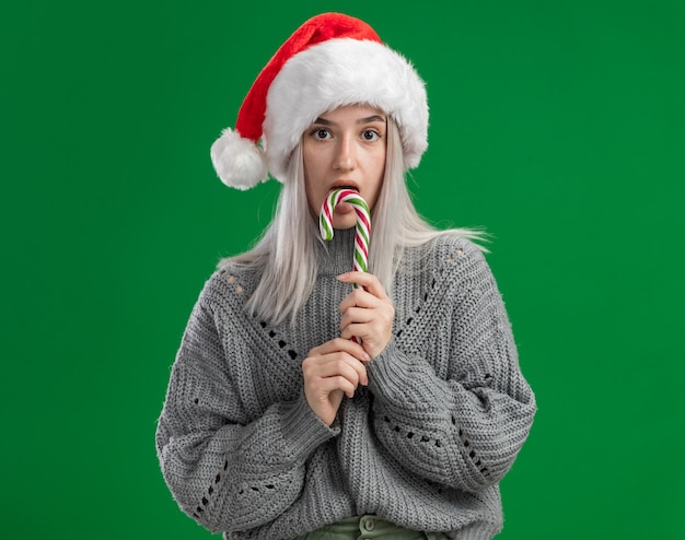 Bezpłatne zdjęcie młoda blond kobieta w zimowym swetrze i santa hat trzyma trzcinę cukrową z poważną twarzą, która spróbuje go stojąc nad zieloną ścianą