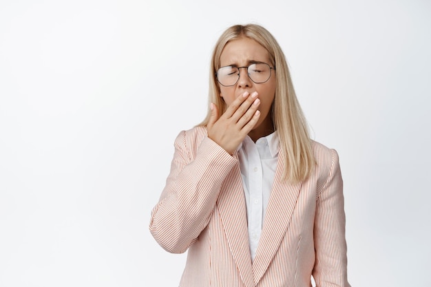 Młoda blond kobieta ziewa w pracy, ubrana w garnitur i okulary zakrywające usta ręką wyczerpaną, mająca zmęczenie białe tło