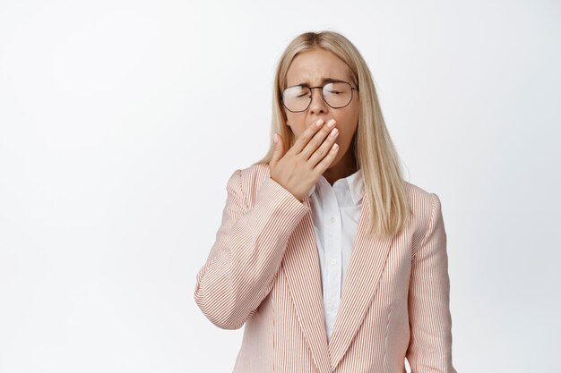 Młoda blond kobieta ziewa w pracy, ubrana w garnitur i okulary zakrywające usta ręką wyczerpaną, mająca zmęczenie białe tło