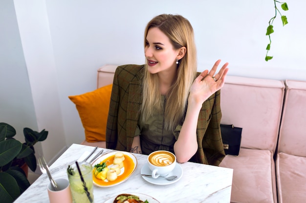 Młoda Blondynka Bardzo Uśmiechnięta Kobieta Delektuje Się Smacznym I Zdrowym Brunchem Z Tostami Z Awokado Z łososia, Cappuccino, Lemoniadą I Deserem, Elegancki Strój, Lekkie, Fantazyjne Wnętrze.