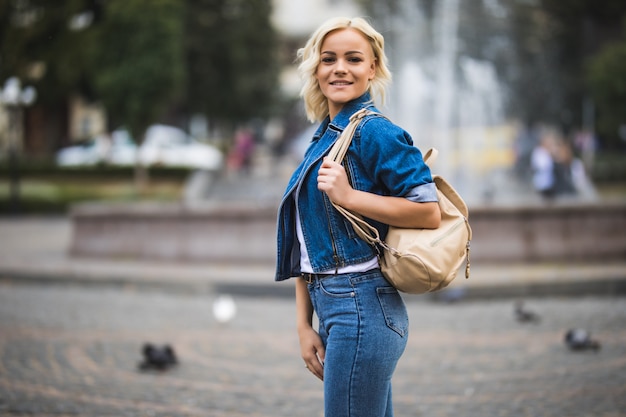 Młoda blondynka na ulicy streetwalk square fontain ubrana w dżinsowy apartament z torbą na ramieniu w słoneczny dzień