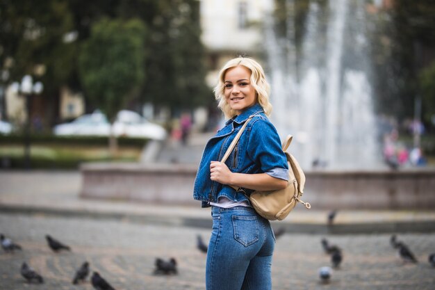 Młoda blondynka na ulicy streetwalk square fontain ubrana w niebieskie dżinsy z torbą na ramieniu w słoneczny dzień