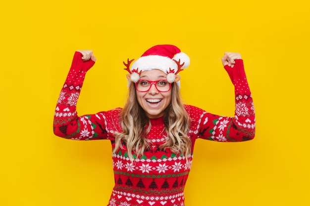 Młoda blondynka w świątecznej czapce mikołaja i swetrze w jelenia jest zadowolona z podniesionymi rękami