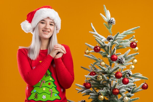 Młoda blondynka w świąteczny sweter i santa hat trzymając się za ręce razem szczęśliwy i wesoły uśmiechnięty
