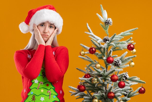 młoda blondynka w świątecznym swetrze i czapce mikołaja patrząc na kamery ze smutnym wyrazem twarzy z rękami na policzkach stojąca obok choinki na pomarańczowym tle