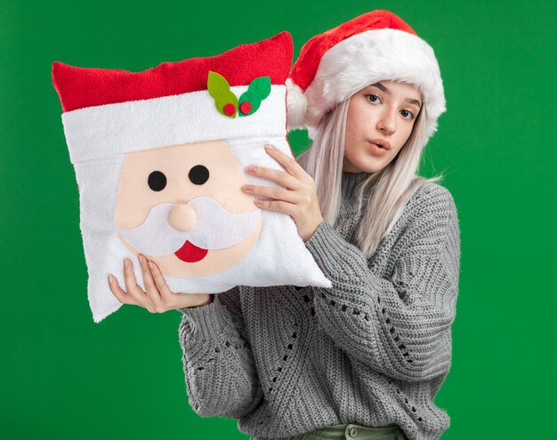 Młoda blondynka w zimowy sweter i santa hat trzyma poduszkę świąteczną patrząc na kamery z pewną siebie wypowiedzi stojącej na zielonym tle