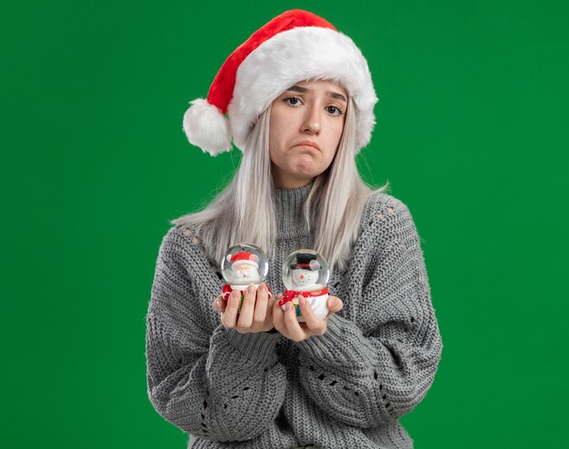 Młoda blondynka w zimowym swetrze i czapce mikołaja trzymająca świąteczne zabawki śnieżne kule patrząc na kamerę zdezorientowana bez odpowiedzi stojąc na zielonym tle