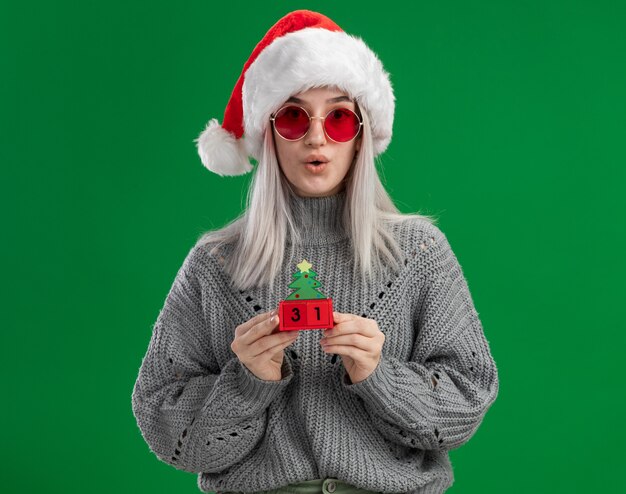 Młoda blondynka w zimowym swetrze i czapce mikołaja w czerwonych okularach trzyma kostki zabawek z datą szczęśliwego nowego roku patrząc na kamerę zaskoczona stojąc na zielonym tle