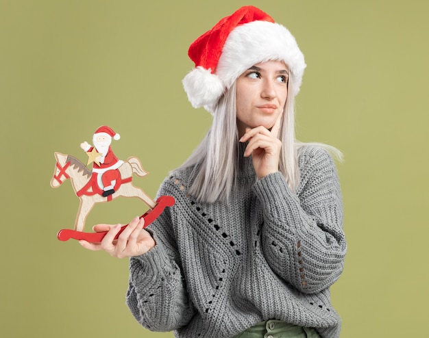 Bezpłatne zdjęcie młoda blondynka w zimowym swetrze i santa hat trzyma świąteczną zabawkę, patrząc na bok, zdziwiona, stojąc nad zieloną ścianą