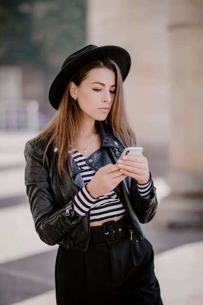 Młoda brązowowłosa dziewczyna w skórzanej kurtce, czarnym kapeluszu na miejskiej promenadzie i gra na telefonie komórkowym