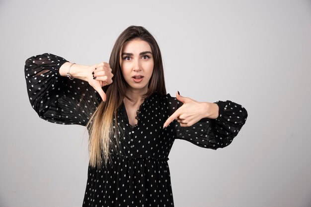 Młoda brunetka kobieta stoi i pokazuje odrzucenie i negatyw z kciukiem w dół gestem.