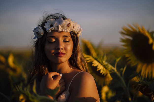Młoda brunetka kobieta w koronie kwiatu z zamkniętymi oczami, ciesząc się słońcem w słonecznikowym polu