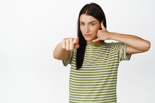 Młoda brunetka kobieta wskazując palcem na aparat pokazujący gest telefonu zadzwoń do nas skontaktuj się z firmą stojącą na białym tle