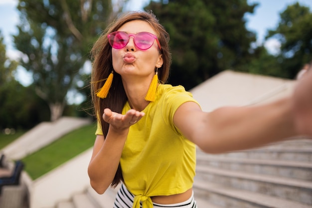 Młoda, całkiem stylowa uśmiechnięta kobieta robi selfie w parku miejskim, pozytywna, emocjonalna, ubrana w żółtą górę, różowe okulary przeciwsłoneczne, trend w modzie na lato, długie włosy, dobra zabawa