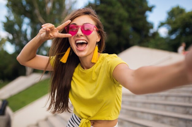 Młoda, całkiem stylowa uśmiechnięta kobieta robi selfie w parku miejskim, pozytywna, emocjonalna, ubrana w żółtą górę, różowe okulary przeciwsłoneczne, trend w modzie na lato, długie włosy, pokazujący znak pokoju