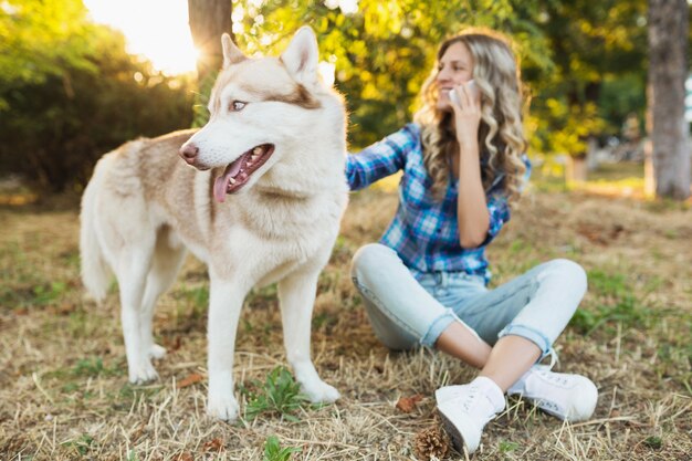 Młoda całkiem uśmiechnięta szczęśliwa blond kobieta bawi się z psem rasy husky w parku w słoneczny letni dzień