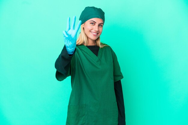 Młoda chirurg urugwajska kobieta w zielonym mundurze na białym tle na niebieskim tle szczęśliwa i licząca trzy palcami