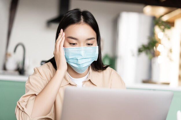 Młoda chora azjatycka dziewczyna w medycznej masce na twarz, korzystająca z laptopa pracującego w domu podczas kwarantanny