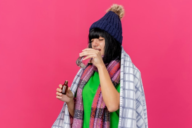 Bezpłatne zdjęcie młoda chora kobieta w czapce zimowej i szaliku owinięta w kratę trzymająca lekarstwo w szklance do picia szklanka wody na różowej ścianie z miejscem na kopię
