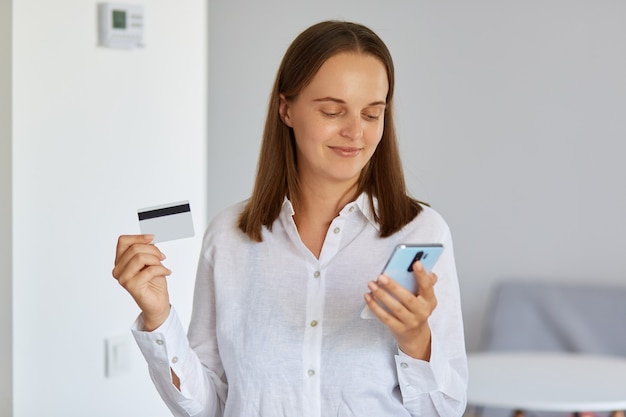 Młoda ciemnowłosa kobieta ubrana w białą koszulę pokazującą kartę kredytową i wprowadzanie danych w inteligentny telefon dla płatności online, patrząc na ekran urządzenia z pozytywnym wyrażeniem.