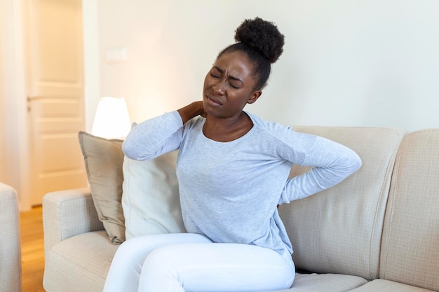 Młoda Czarna kobieta cierpiąca na ból pleców w domu Portret młodej dziewczyny siedzącej na kanapie w domu z bólem głowy i pleców Piękna kobieta mająca ból kręgosłupa lub nerek