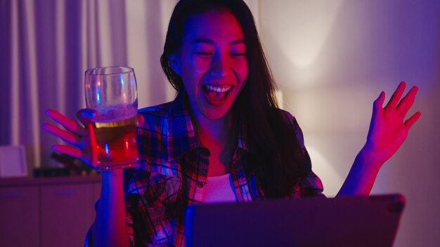 Młoda dama Azji pije piwo, bawiąc się szczęśliwy moment disco neon night party wydarzenie online celebracja za pośrednictwem wideorozmowy w salonie w domu.