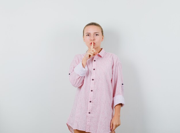 Młoda dama pokazuje gest ciszy w różowej koszuli i wygląda elegancko
