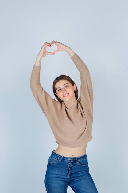 Bezpłatne zdjęcie młoda dama w beżowym swetrze, dżinsach pokazujących gest serca i wyglądających ładnie, widok z przodu.