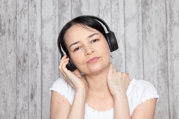 młoda dama w białej koszuli słuchania muzyki w czarnych słuchawkach na szarej ścianie