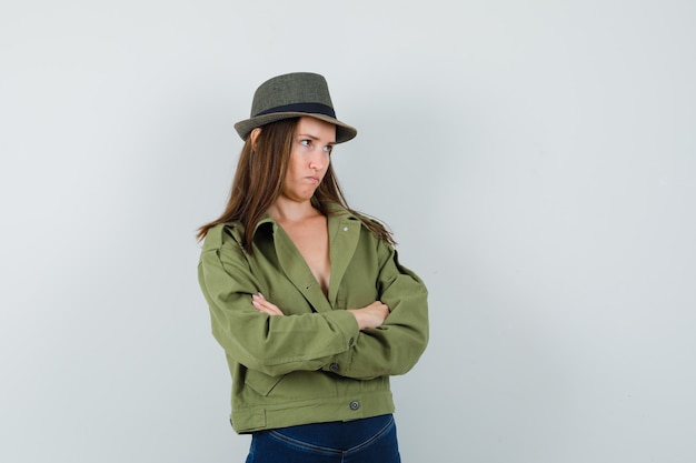 Młoda dama w kapeluszu spodnie kurtka stojąca ze skrzyżowanymi rękami i wyglądająca ponuro