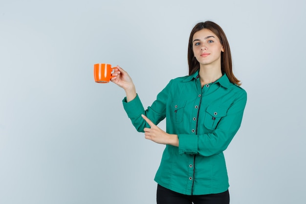 Młoda dama w koszuli trzymająca pomarańczową filiżankę herbaty, wskazująca na lewy górny róg i wyglądająca na pewną siebie, widok z przodu.