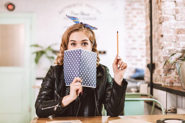 Młoda dama w skórzanej kurtce siedzi przy stole z ołówkiem w dłoni i ze zdumieniem patrzy w kamerę, zakrywając twarz notatnikiem w kawiarni
