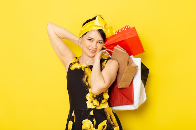 Bezpłatne zdjęcie młoda dama w widoku z przodu, w żółto-czarnym kwiatowym stroju z żółtym bandażem na głowie, trzymająca paczki z zakupami na żółtym
