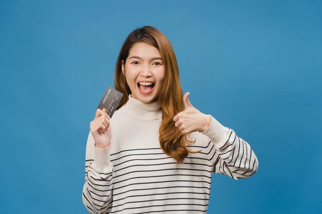 Młoda dama z Azji pokazuje kartę kredytową z pozytywnym wyrazem twarzy, uśmiecha się szeroko, ubrana w zwykłe ubranie, czując szczęście i stojąc na białym tle na niebieskiej ścianie