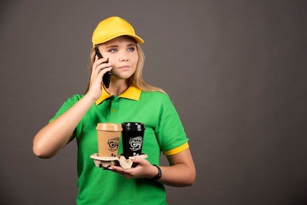 Bezpłatne zdjęcie młoda deliverywoman z filiżankami kawy, mówiąc na smartfonie.