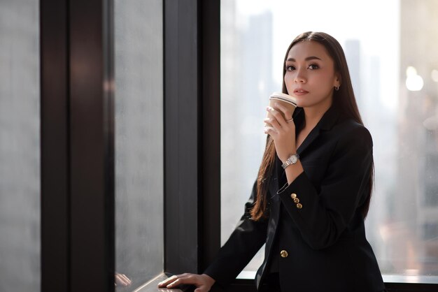 Młoda dorosła inteligentna azjatycka kobieta biznesu w czarnym garniturze dorywczo trzymająca filiżankę kawy w czasie przerwy
