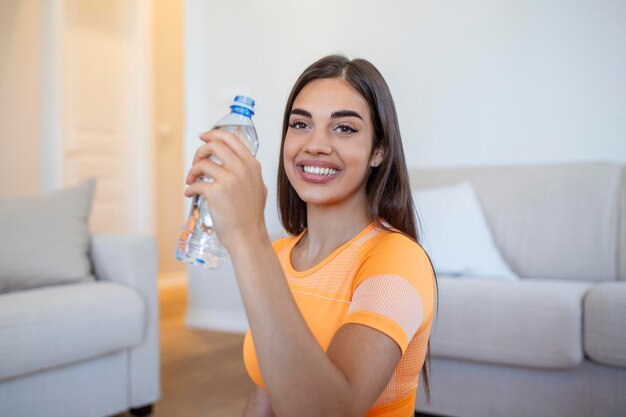 Młoda dorosła kobieta wody pitnej z plastikowej butelki siedzi na macie fitness i odpoczywa po treningu w domu