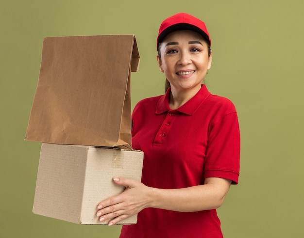 Młoda dostawa kobieta w czerwonym mundurze i czapce trzymająca papierowe opakowanie i kartonowe pudełko szczęśliwa i pozytywnie uśmiechnięta stojąca nad zieloną ścianą