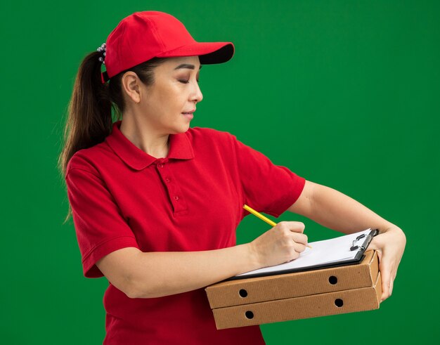 Młoda dostawa kobieta w czerwonym mundurze i czapce, trzymająca pudełka po pizzy i schowek z długopisem, pisząca coś z pewną miną