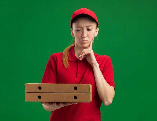 Młoda dostawcza dziewczyna w czerwonym mundurze i czapce trzymająca pudełka po pizzy, patrząca na nie zdziwiona palcem na brodzie, stojąca nad zieloną ścianą