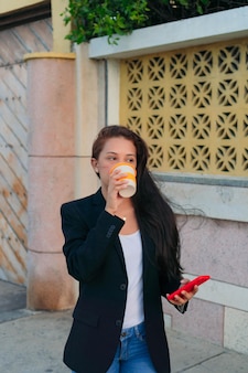 Młoda dyrektorka pijąca kawę i trzymająca telefon komórkowy, stojąc w wolnym czasie