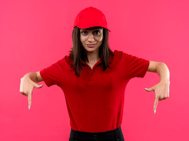 Młoda dziewczyna dostawy ubrana w czerwoną koszulkę polo i czapkę wskazującą palcami wskazującymi w dół, uśmiechnięta pewnie stojąca nad różową ścianą