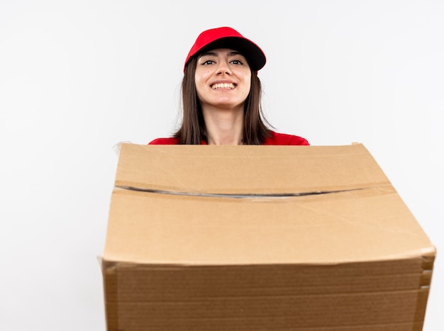 Młoda dziewczyna dostawy ubrana w czerwony mundur i czapkę, trzymając duży karton z uśmiechem na twarzy stojącej nad białą ścianą