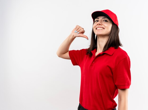 Młoda dziewczyna dostawy ubrana w czerwony mundur i czapkę, wskazując na siebie, uśmiechając się pewnie stojąc na białej ścianie