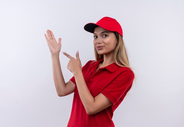 młoda dziewczyna dostawy ubrana w czerwony mundur i czapkę wskazuje jej rękę na białym tle na białej ścianie