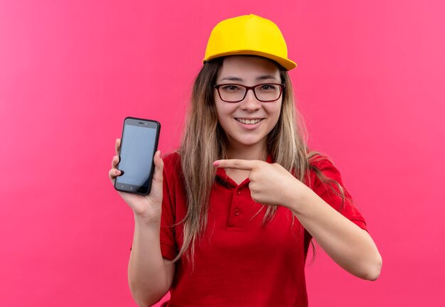 Młoda dziewczyna dostawy w czerwonej koszulce polo i żółtej czapce pokazuje smartfon, wskazując palcem wskazującym na to uśmiecha się