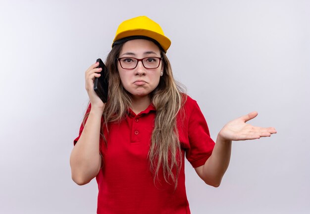 Młoda dziewczyna dostawy w czerwonej koszulce polo i żółtej czapce rozmawia przez telefon komórkowy, patrząc zdezorientowany wzruszając ramionami